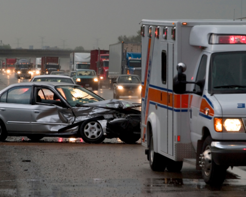 A heavily damaged car sits at a crash scene with an ambulance nearby and traffic in the background, illustrating the serious impact of a drunk driving accident and the importance of pursuing compensation under Texas DUI injury laws